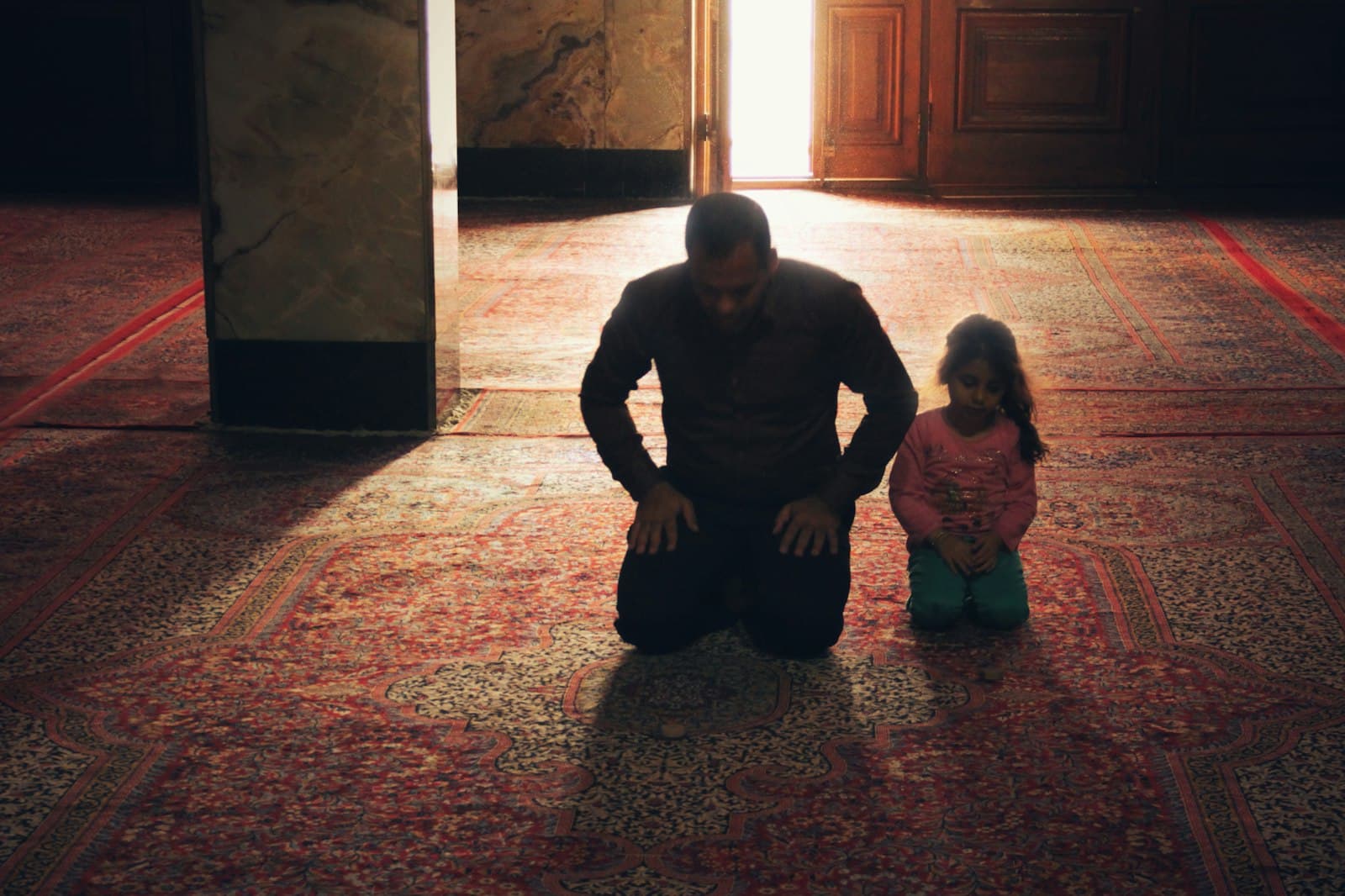 A father and young daughter praying together in a mosque lit by sunlight