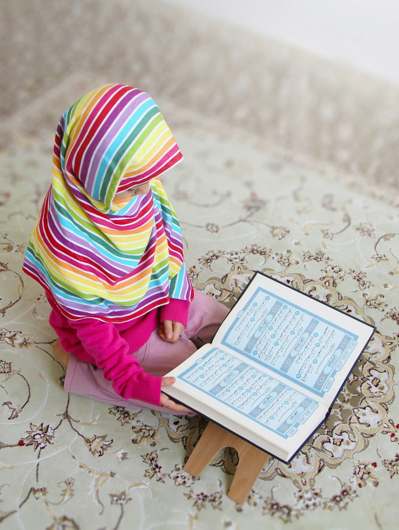 A young girl in a colorful hijab reading the Quran on a wooden stand at home